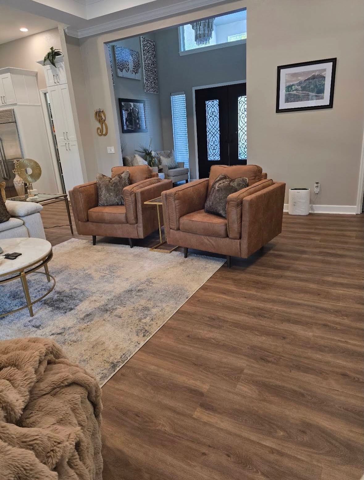 Modern living room with brown chairs, patterned rug, wood flooring, and a front entryway.