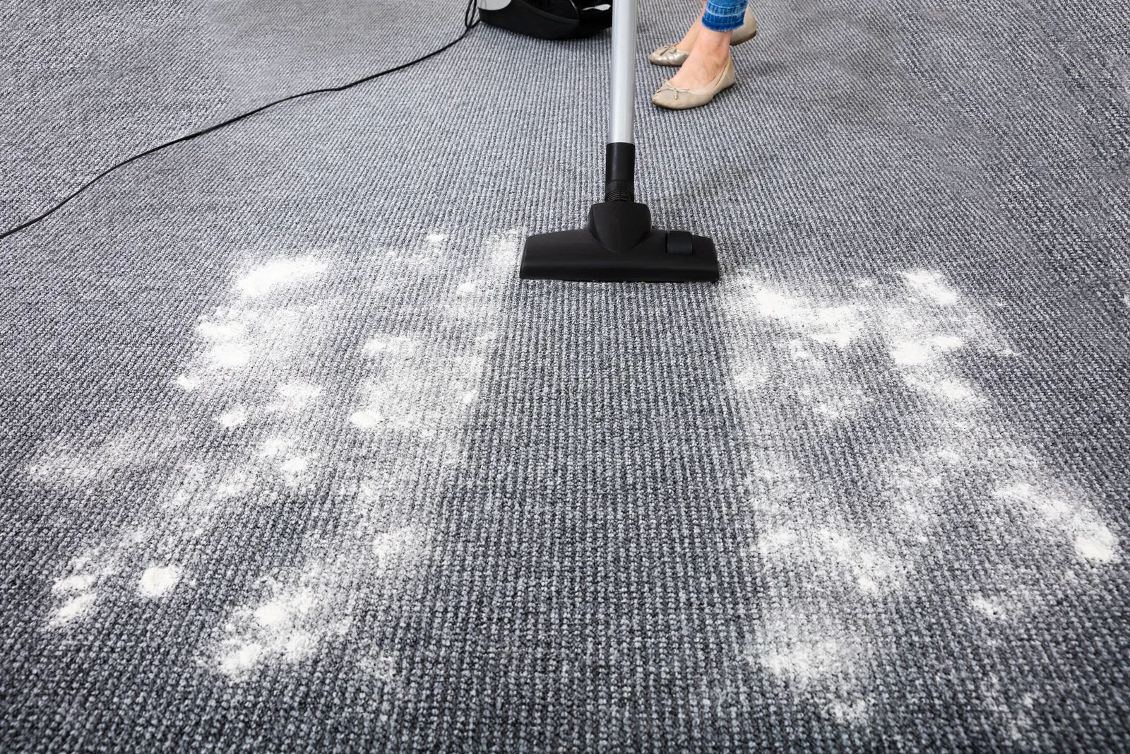 Vacuum cleaner on gray carpet with bright sunlight patches and a person’s foot in the background