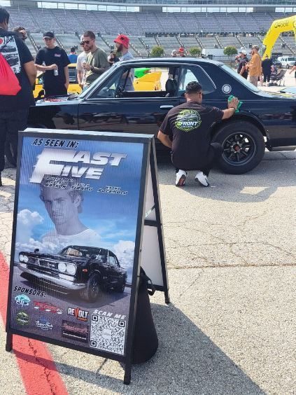 Poster sign in foreground at a car show, with a black classic car and people in the background.