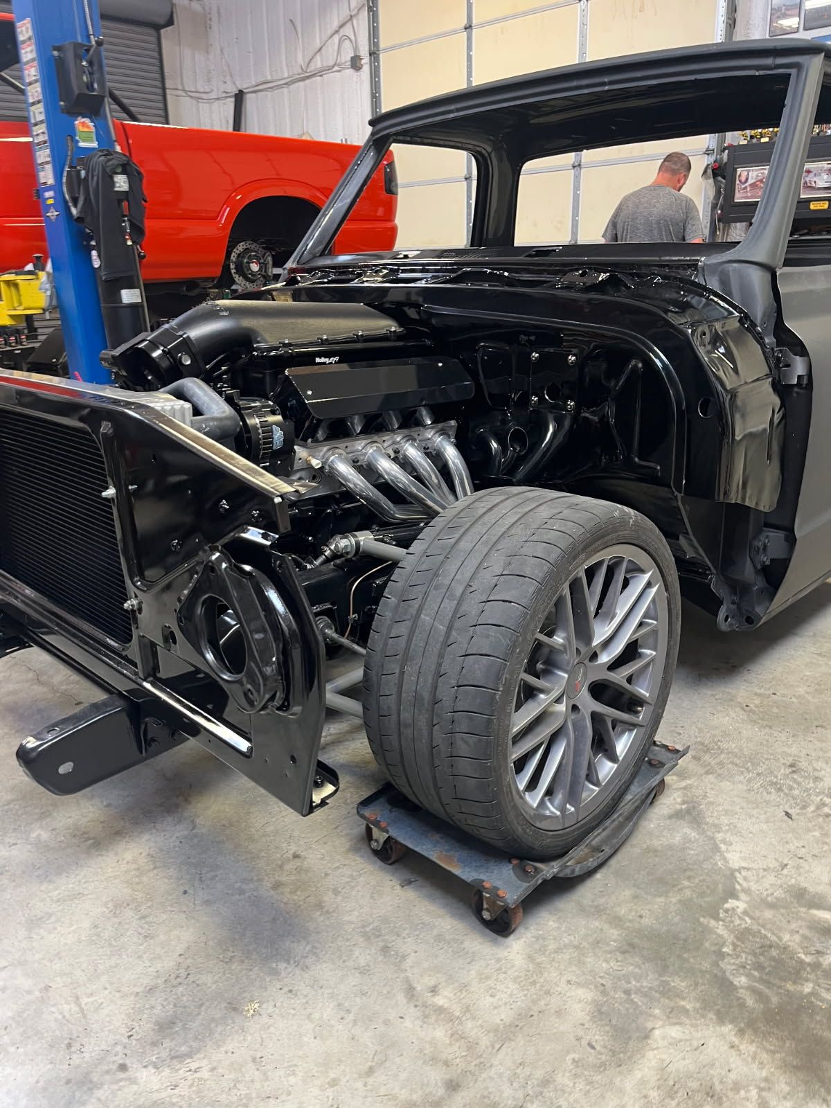 Black hot rod chassis with exposed engine and front wheels in an auto shop, red car and lift in background