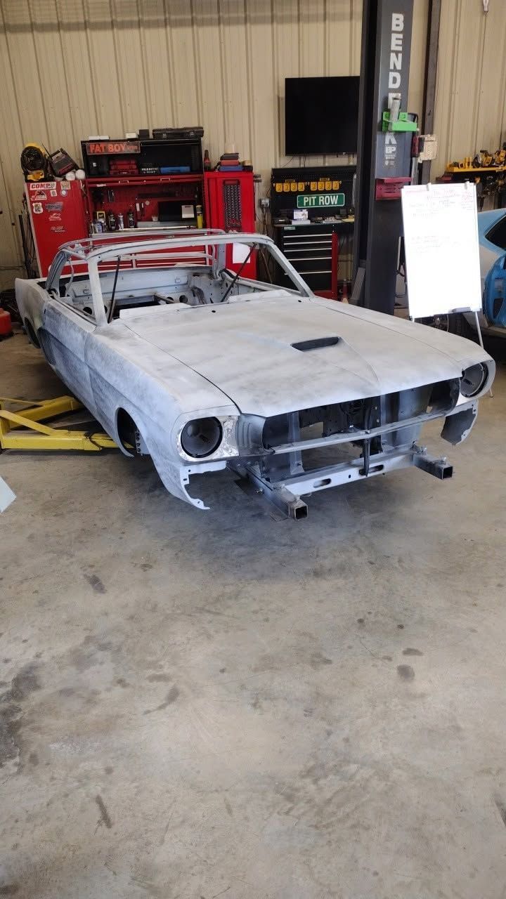 Primer gray classic car shell in a garage, missing front grille and headlights, with red tools in back.