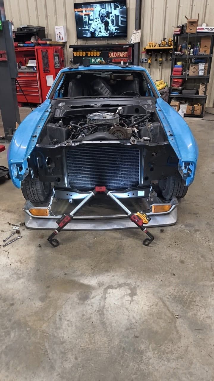 Blue car shell on jack stands in an auto shop, with front panels removed and engine bay exposed