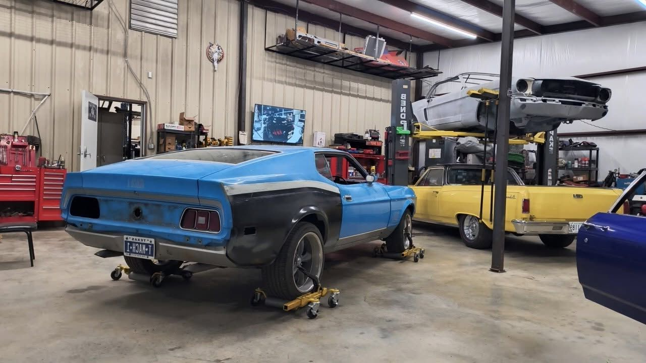 Blue classic car in a repair shop, raised on jack stands with a yellow car and lift in the background