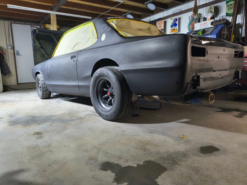 Matte black classic coupe in a garage, with yellow-taped windows and exposed rear wheels.