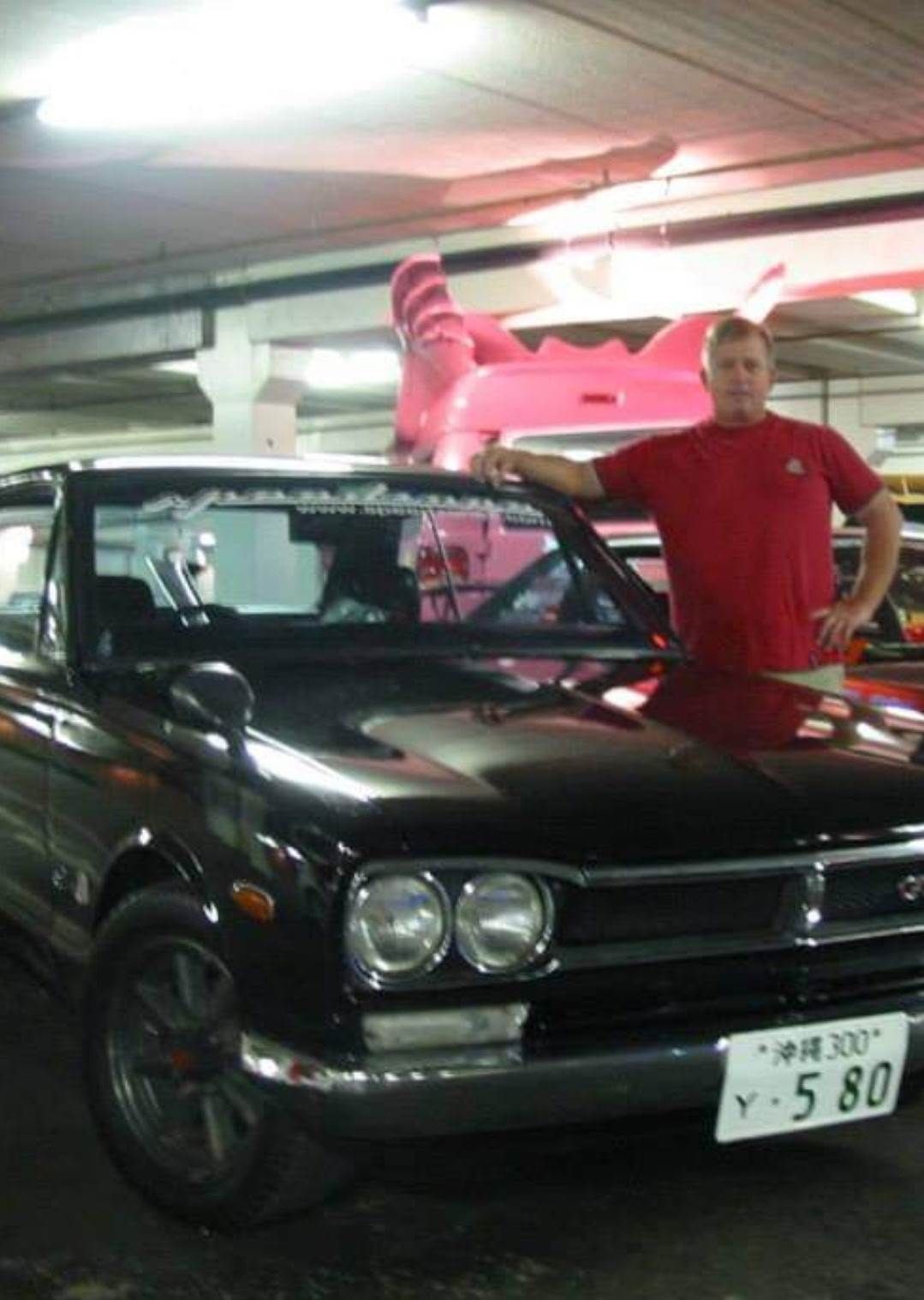 Man standing beside a black classic car in a parking garage, wearing a red shirt.