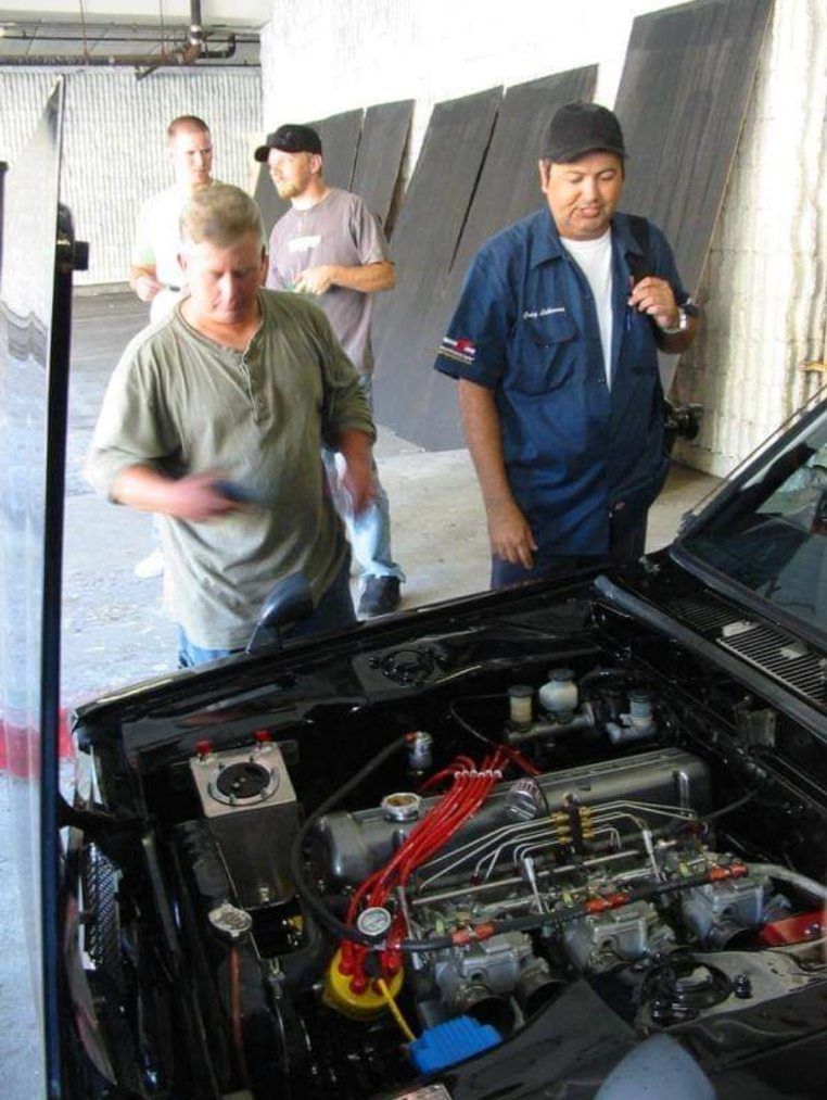 People examining a car engine in a garage, with the hood open and tools nearby.