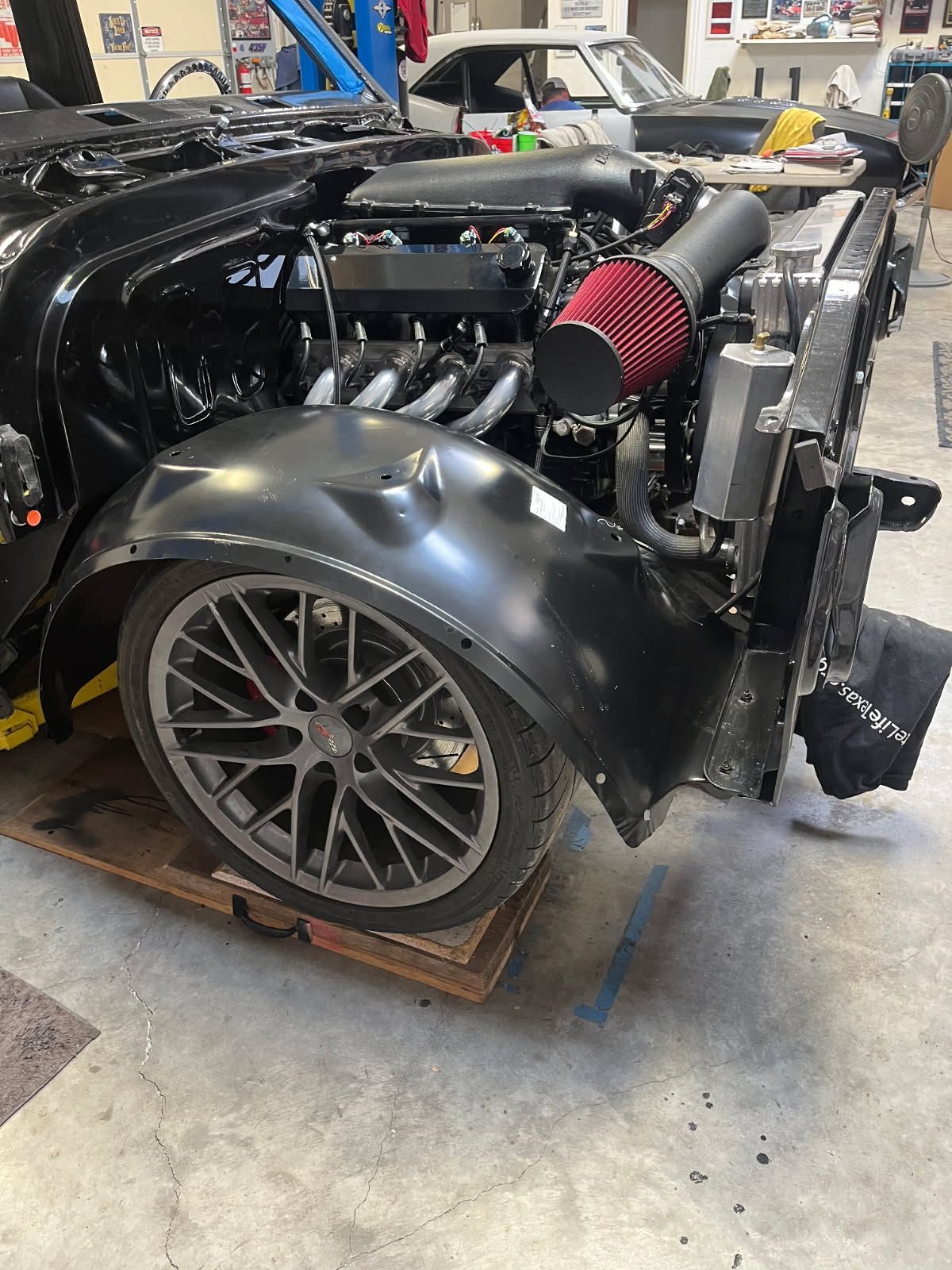 Black race car chassis with exposed engine and red air filter in a workshop, rear wheel visible