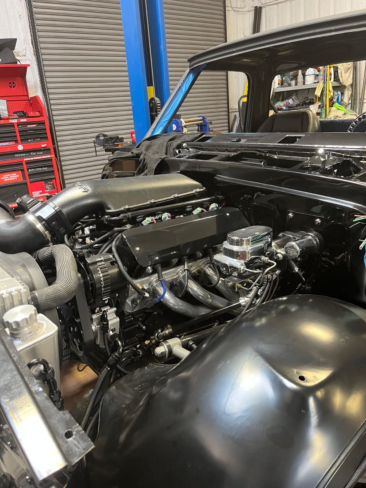 Classic car engine bay with polished chrome components in a workshop, black fenders visible.