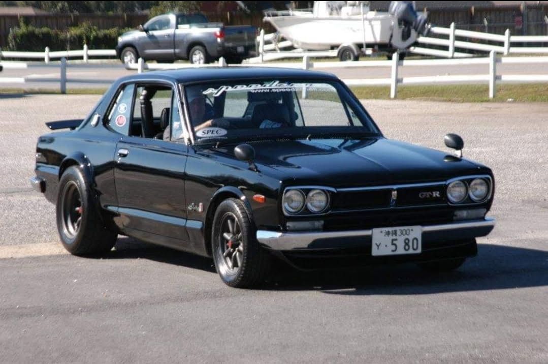 Black classic sedan parked outdoors on a paved lot, with boats and a fence in the background
