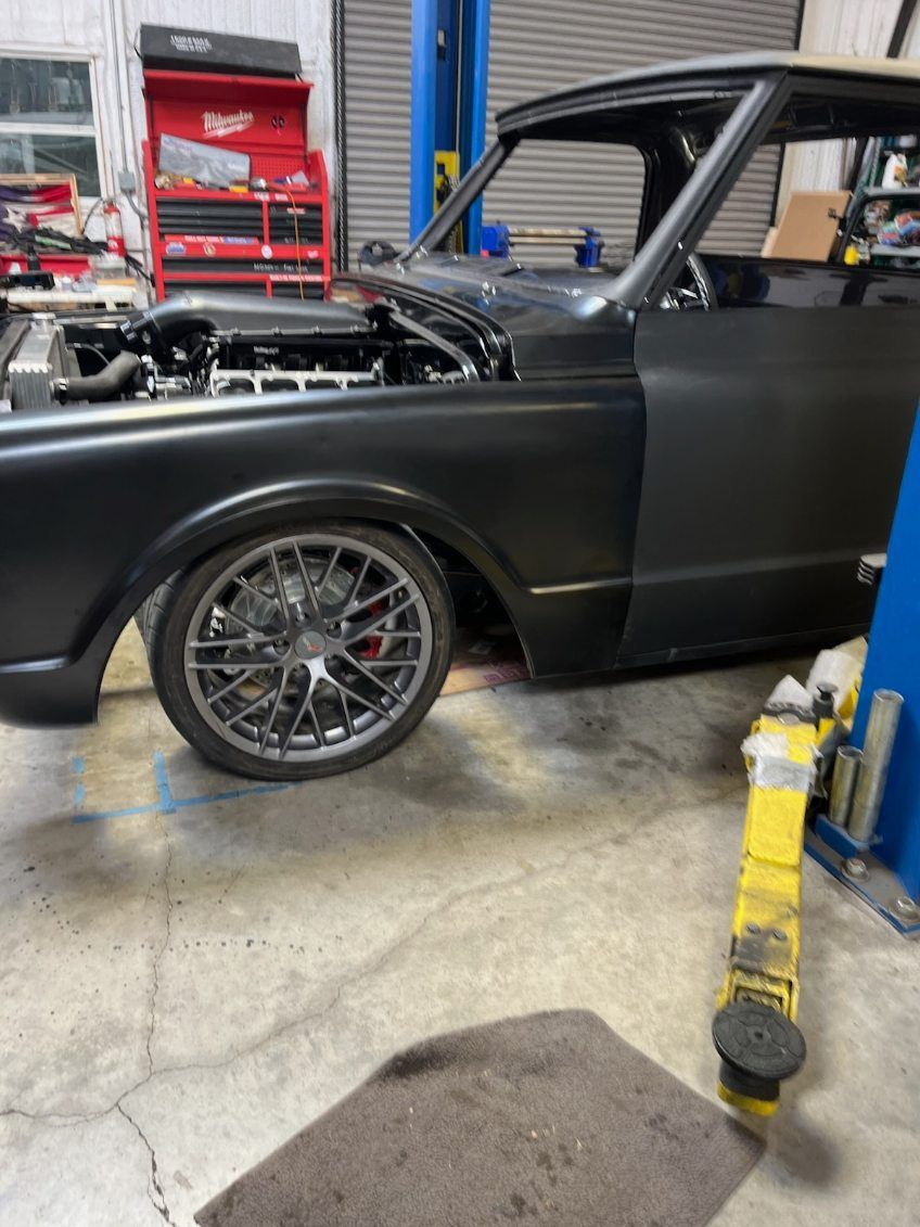 Matte black classic car in an auto shop with hood open and a yellow jack stand nearby