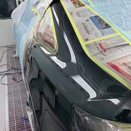 Black car panel in a paint booth, masked with newspaper and yellow tape.