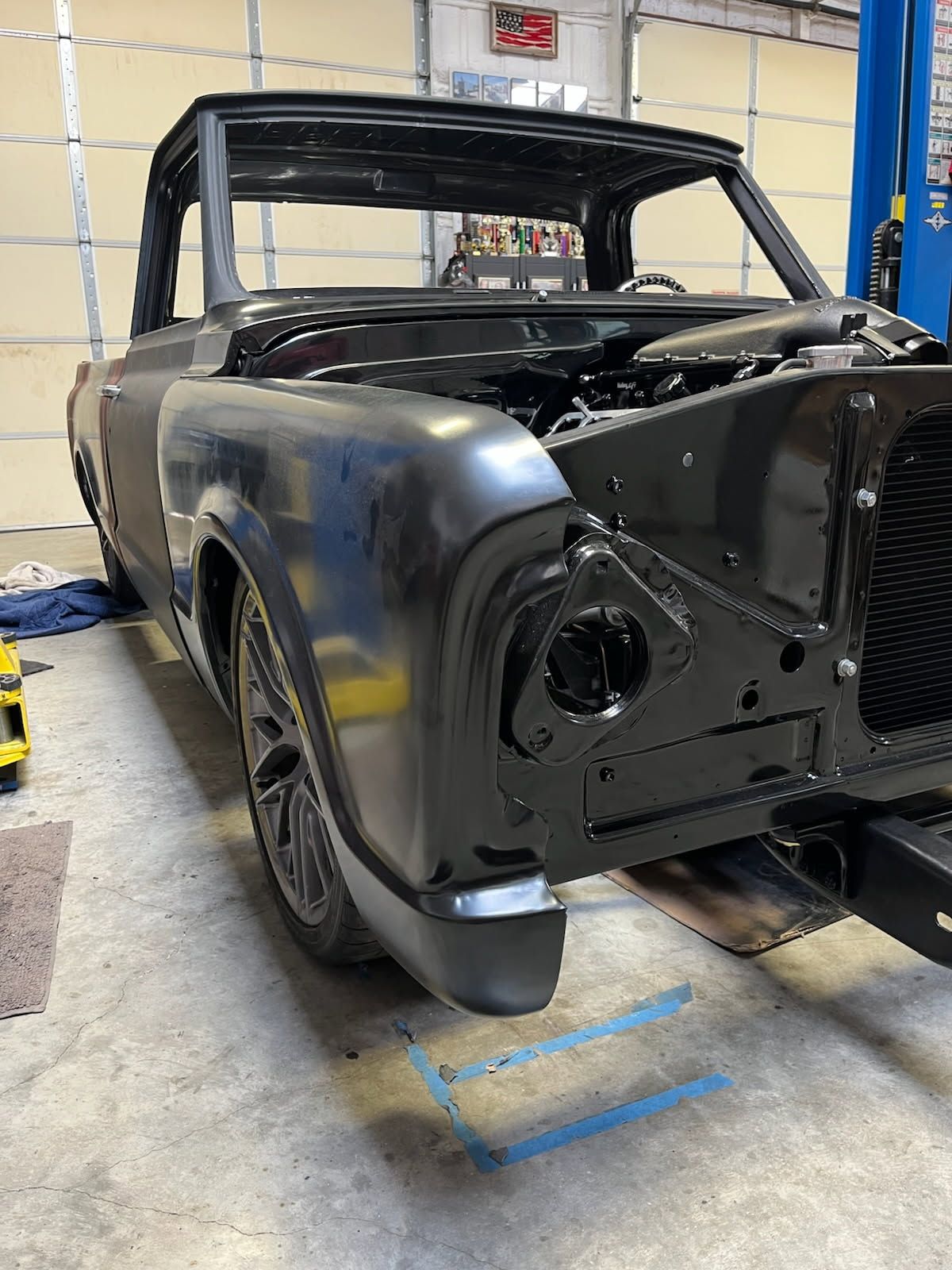 Matte black vintage car body in a garage, missing front grille and headlights, on a lift bay floor.