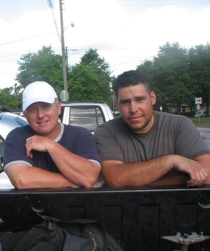 Two men leaning on a black truck tailgate outdoors, one in a white cap and one in a gray shirt.