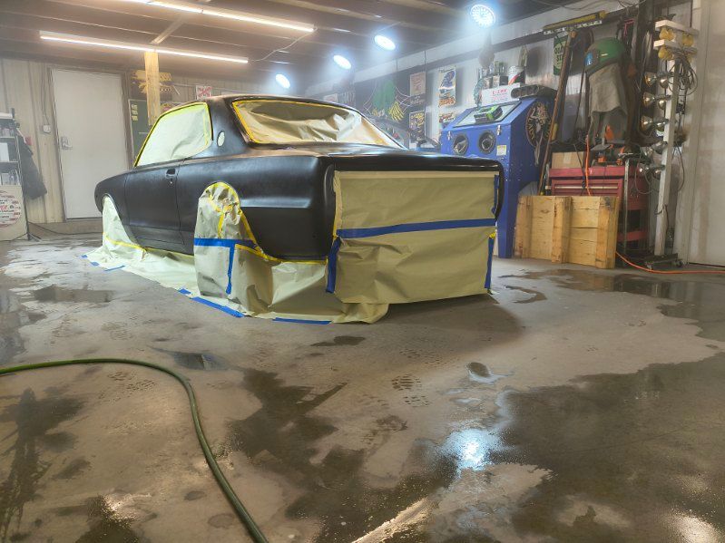 Car in garage covered with masking paper and tape, mid-paint restoration, with wet concrete floor.