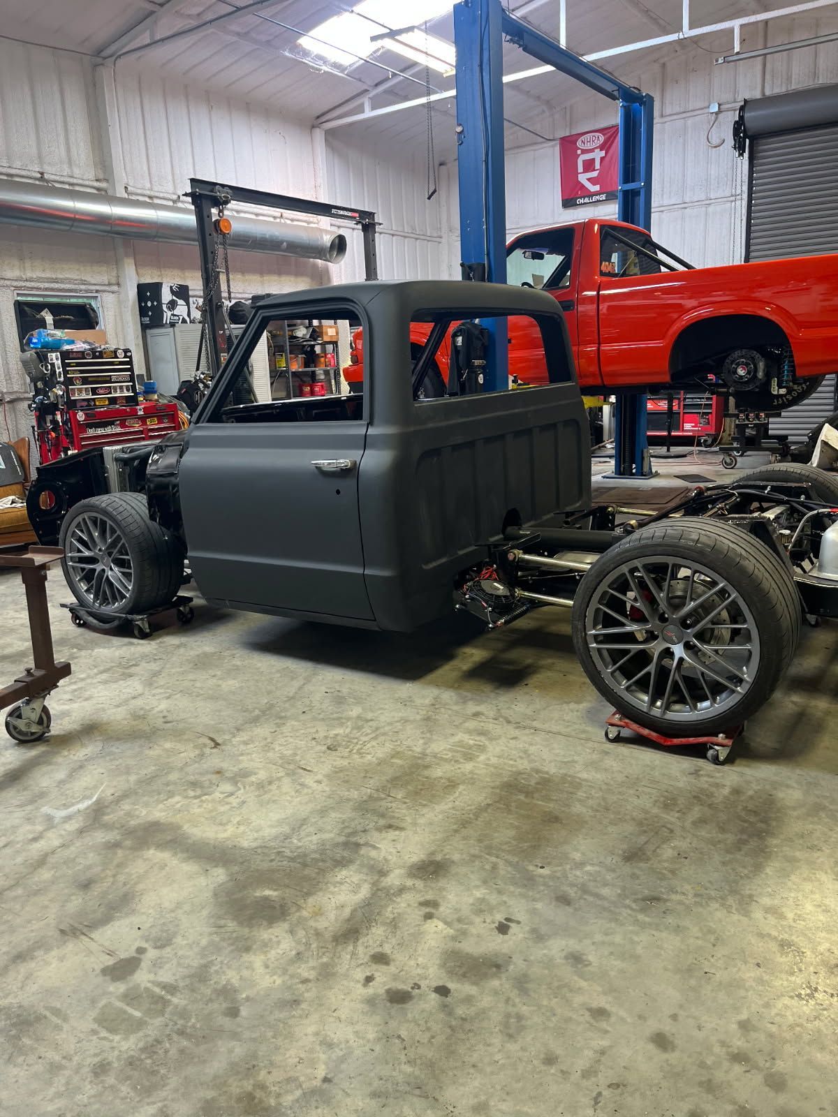 Custom matte-black pickup truck in an auto shop, with a red truck on a lift in the background.