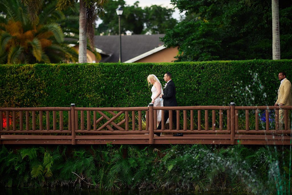 Couple on a wooden bridge, a lush green hedge and pond behind them; man in suit, woman in white dress.