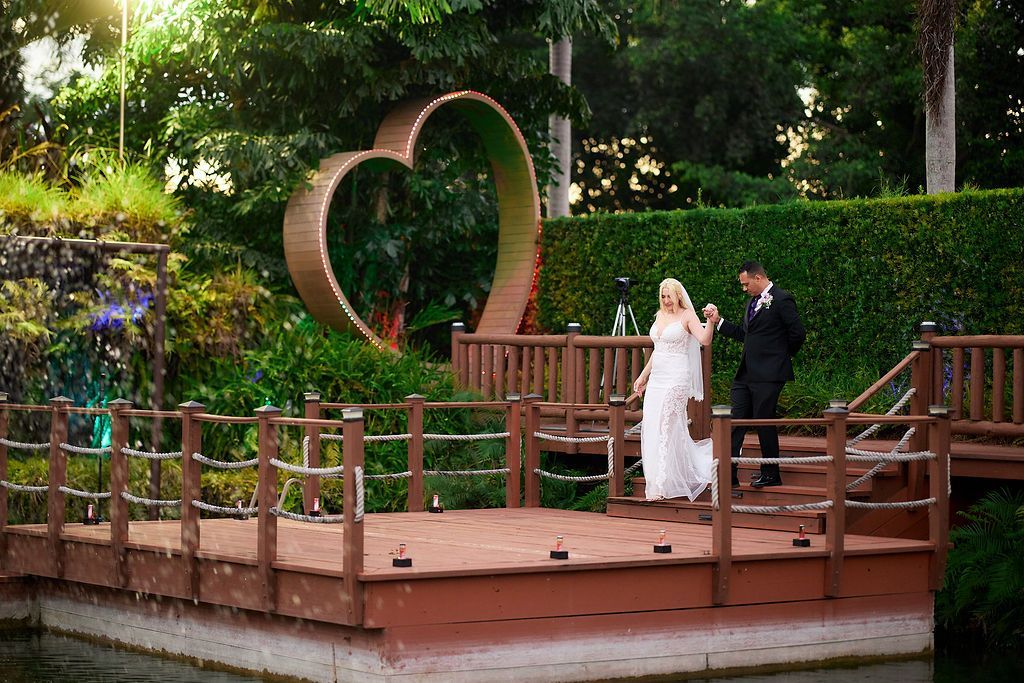 Bride and groom on a wooden platform, walking towards a heart-shaped structure. Outdoor setting with greenery.