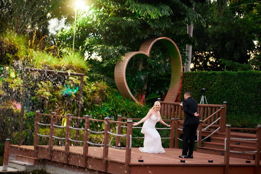 Bride and groom on wooden platform near heart-shaped sculpture. Bride in white gown smiles, groom in suit holds her.