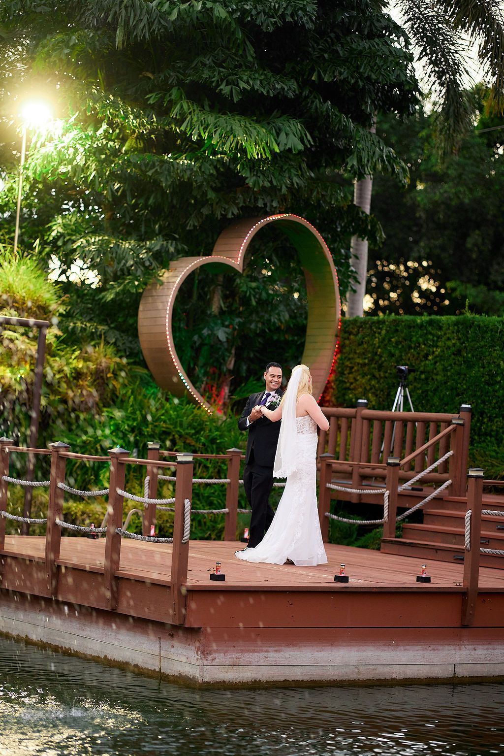 Couple getting married on a wooden deck with a heart-shaped backdrop in a lush outdoor setting.