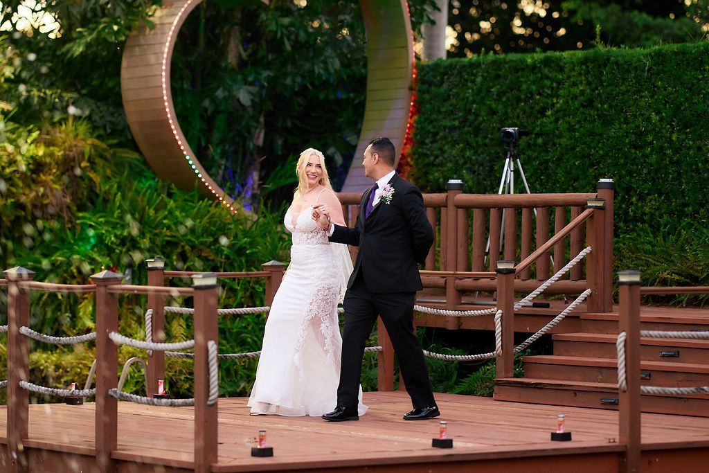 Bride and groom dance on a wooden deck during an outdoor wedding ceremony; heart-shaped arch in background.