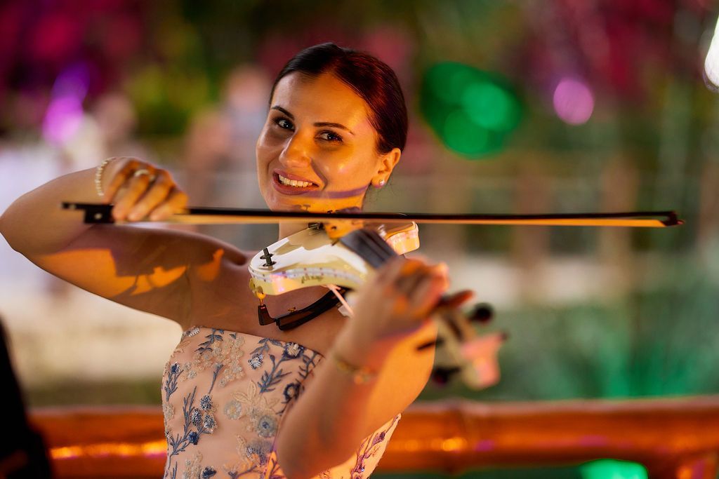 Woman smiles while playing a violin outdoors at night. Bright lights and a festive atmosphere.