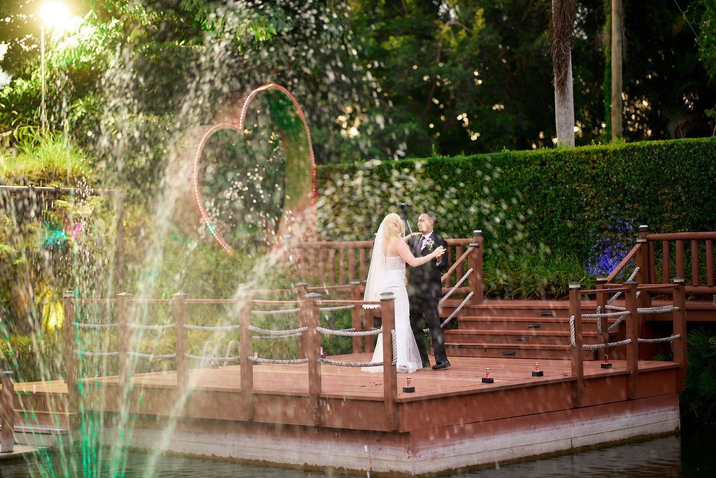 Couple dancing on a wooden stage with heart backdrop, water fountain in the foreground, and lush greenery.