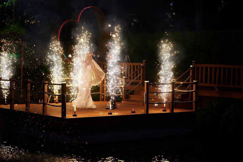 Bride in a white gown stands on a platform with fireworks as part of a wedding celebration.