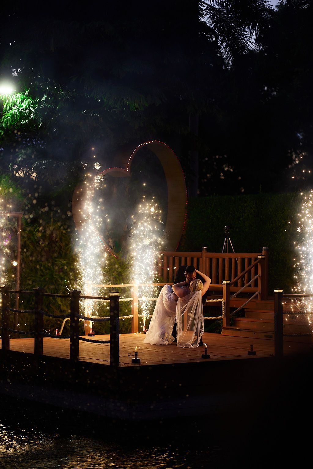 Couple kissing on a wooden stage with sparklers and heart-shaped arch at night.