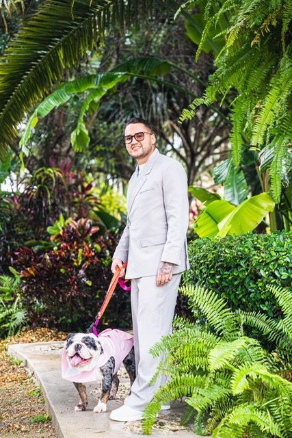 Man in suit walks a bulldog in a pink harness on a leash, among lush green plants.