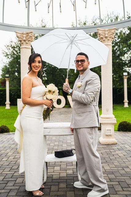 Bride and groom under umbrella at outdoor wedding. Smiling, white dress and suit. 