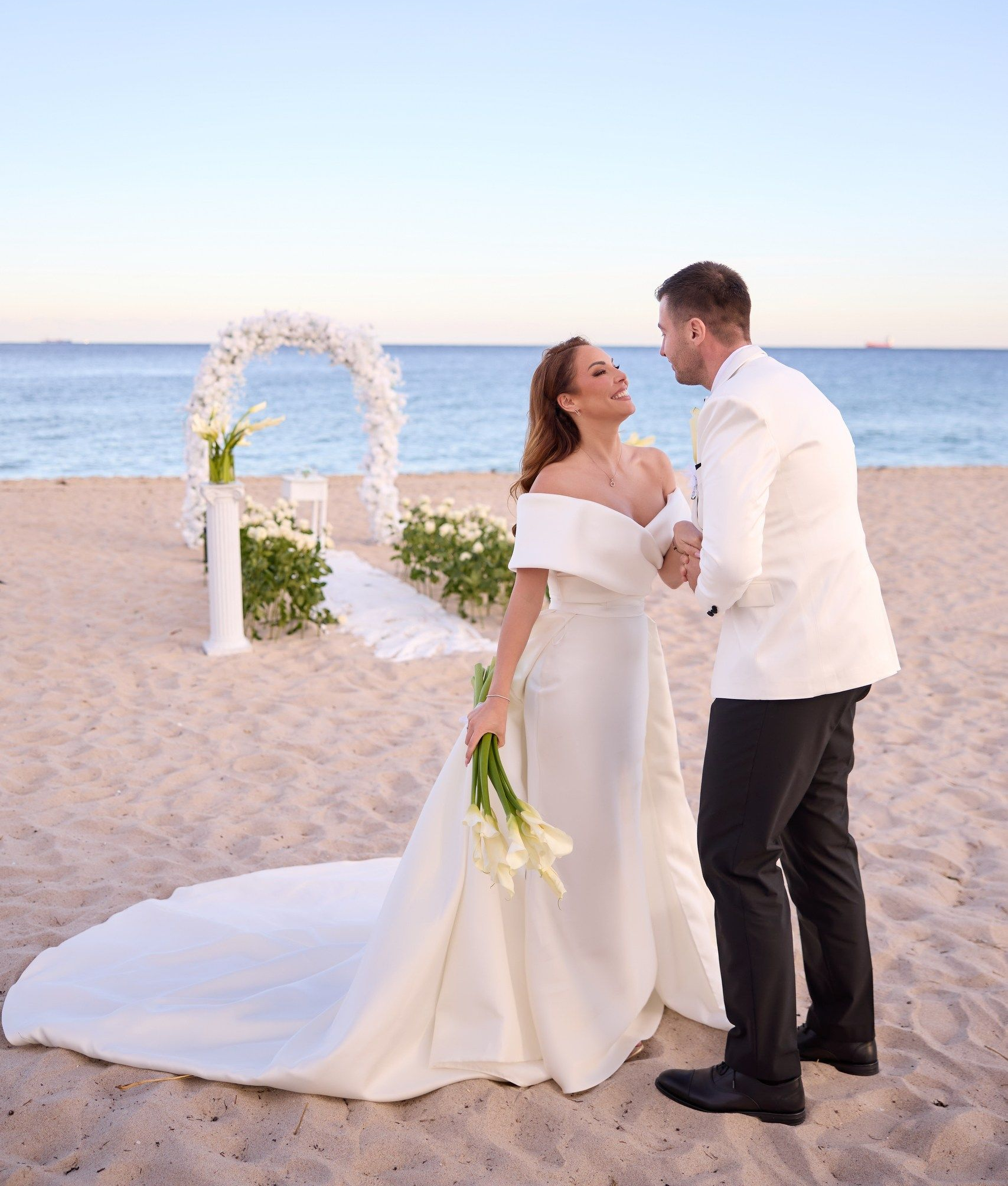 bride and groom on the beach, with white floral wedding arch behind them, looking at each other and smiling