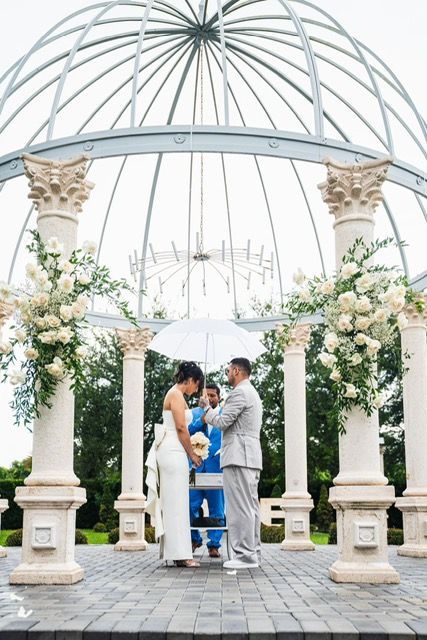 Wedding ceremony under a gazebo. Bride and groom stand with officiant; it’s raining. White floral arrangements on pillars.