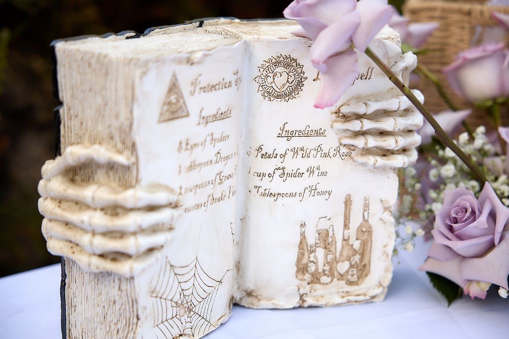 Book-shaped cake, pages open, with carved skeleton hand, faux text, spiderweb design, and purple roses.