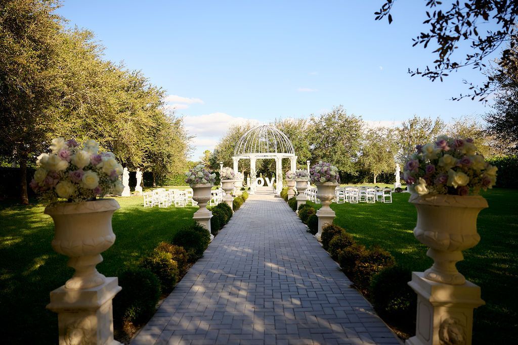 Stone pathway leading to wedding altar in a garden. Floral arrangements in urns line the path.