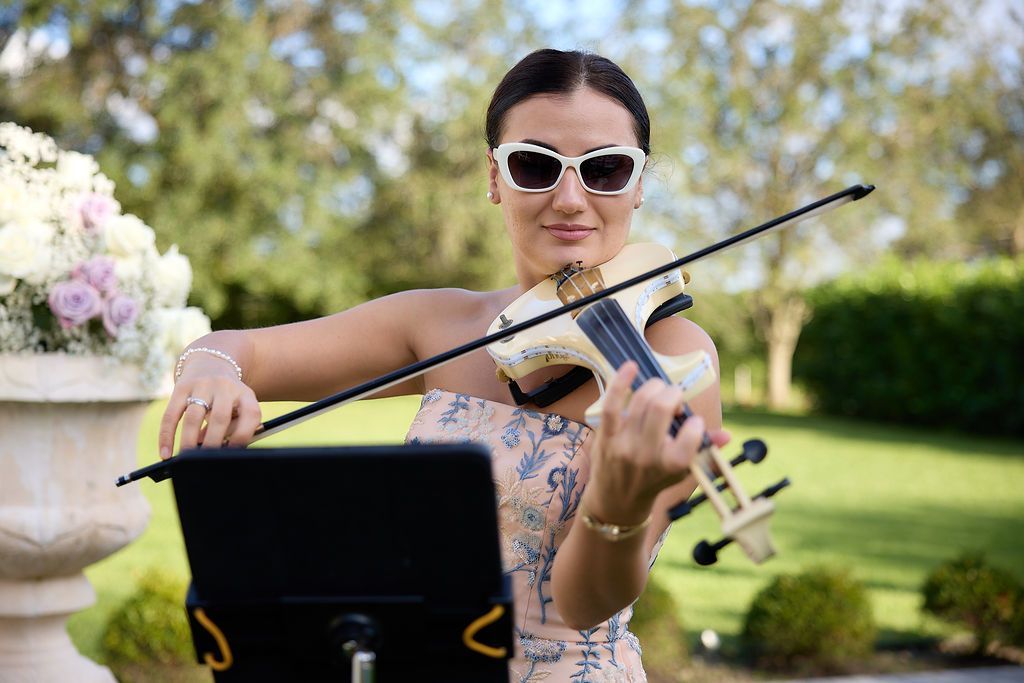 Woman playing a violin outdoors, wearing sunglasses. Flowers and greenery in the background.