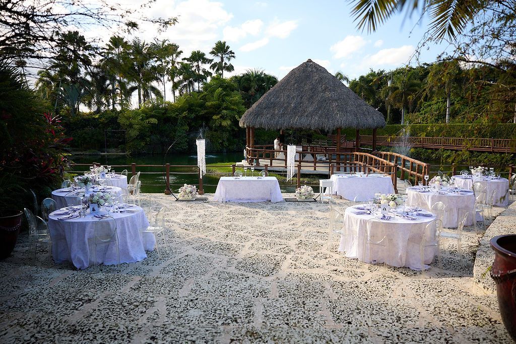 Wedding reception setting with white tables, a thatched roof gazebo, and confetti on the ground, outdoors.