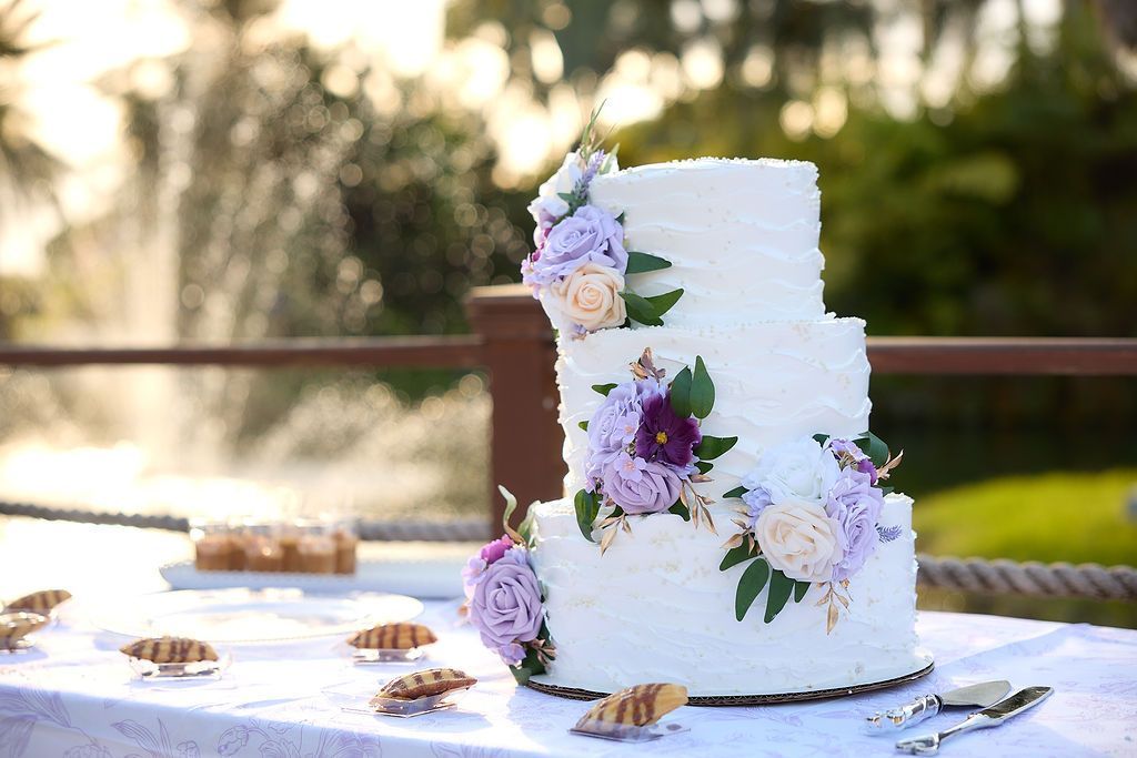 Three-tiered white wedding cake with purple and peach flowers, set outdoors with a fountain in the background.