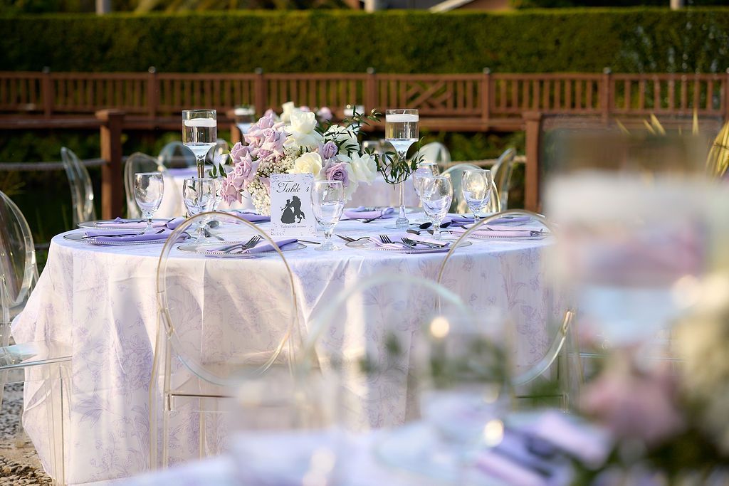 Round table set for an outdoor event with white tablecloth, purple and white flowers, and clear chairs.