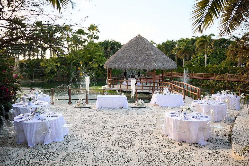 Outdoor wedding reception setup with tables on white ground, thatched gazebo, and greenery.