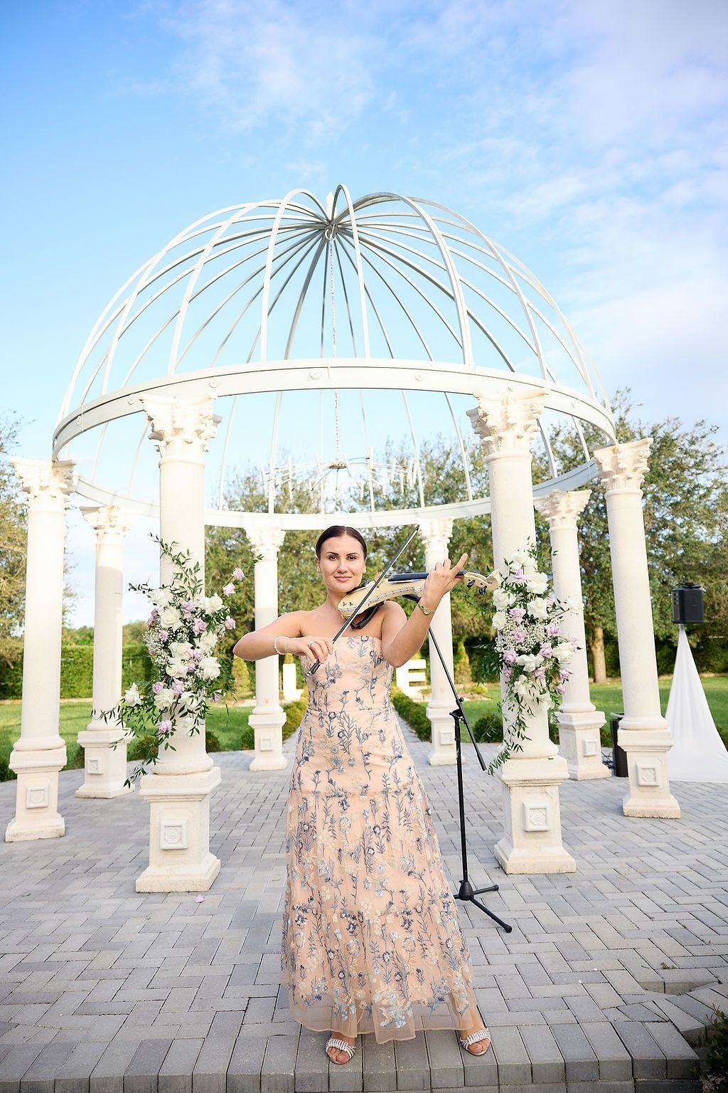 Woman playing violin in front of a gazebo decorated with flowers. She wears a floral dress and stands outdoors.