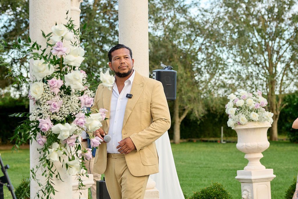 Man in tan suit speaks outdoors near floral arrangement, white pillars, and green lawn.
