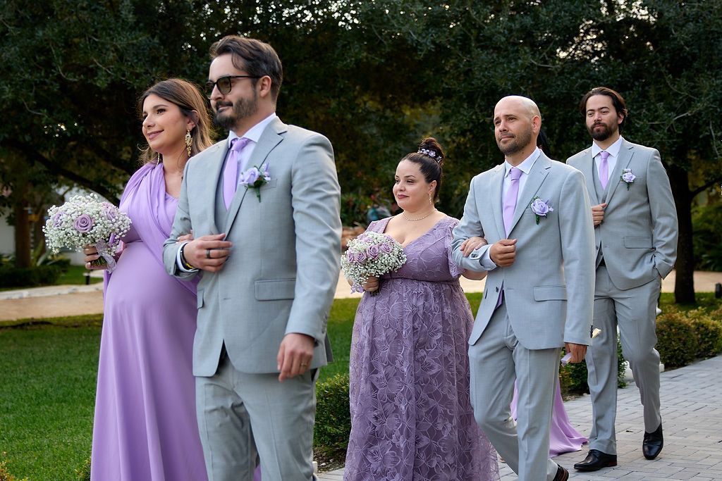 Wedding party in formal attire walking outside, holding bouquets. Lavender and gray color scheme.