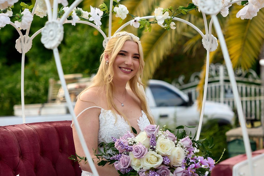 Bride in a white wedding dress holds a bouquet, smiling in front of a decorative archway and bench.