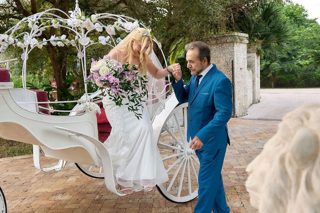 Man in blue suit kissing bride's hand exiting a white horse-drawn carriage at a wedding.