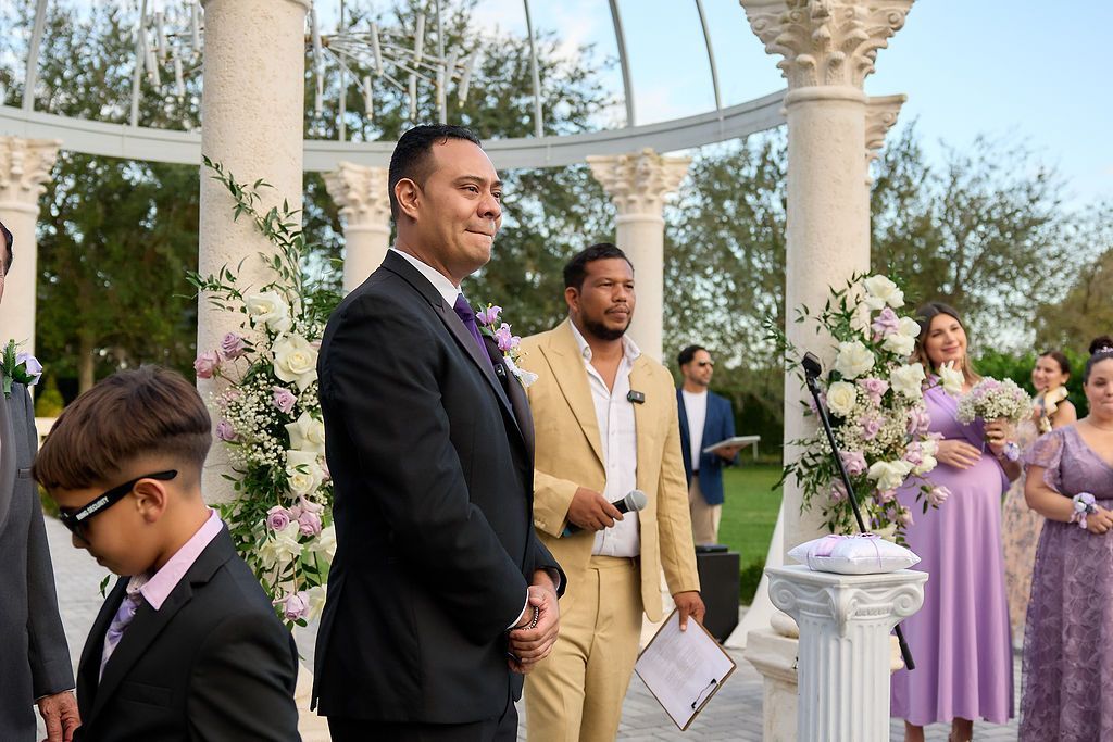 Wedding ceremony outdoors; groom in black suit smiles, officiant speaks into microphone.