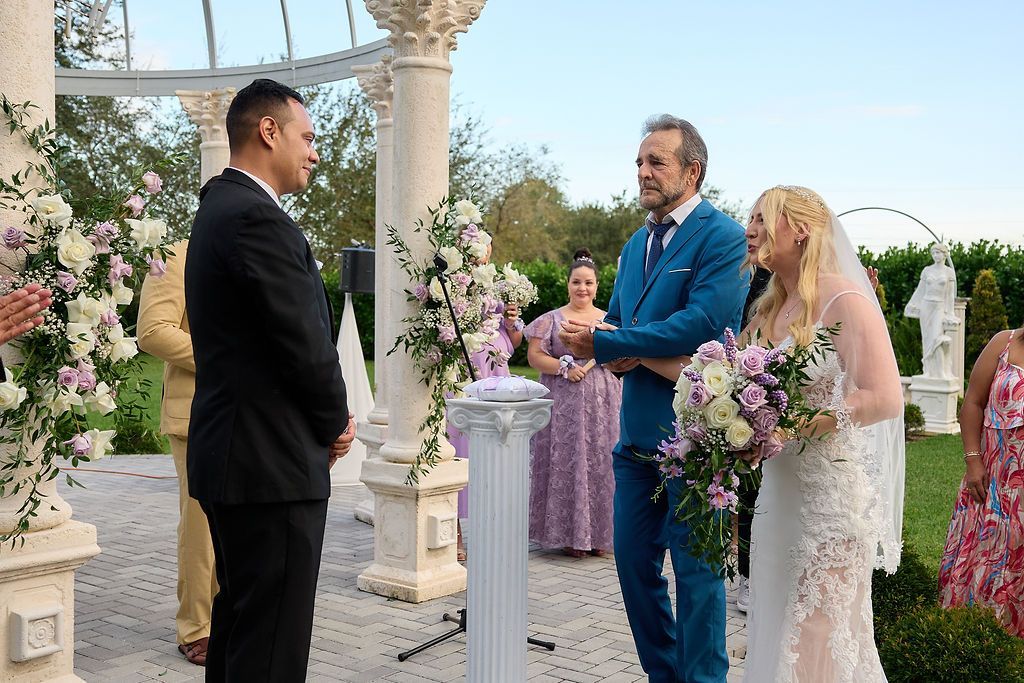 Wedding ceremony. Groom facing officiant and bride under ornate structure. Bride holds flowers. Others watch.