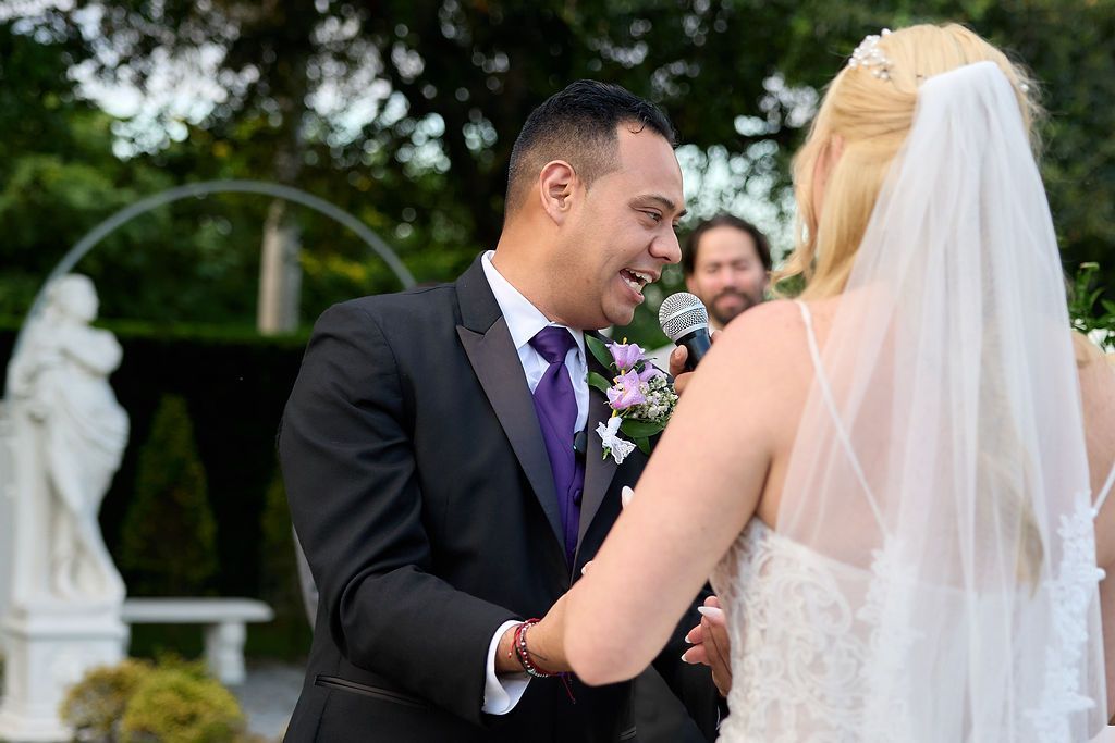 Groom and bride at wedding ceremony. Groom smiles at bride as he holds her hand, with an officiant in the background.