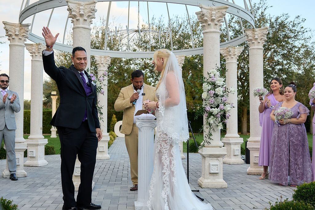 Wedding ceremony: Groom waving, bride with veil, officiant, bridesmaids in lavender dresses, outdoor venue.