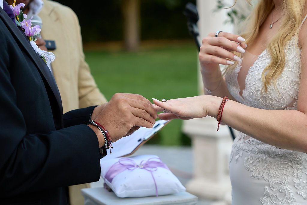 Groom placing a ring on bride's finger during a wedding ceremony. White dress, lavender pillow, outdoor setting.