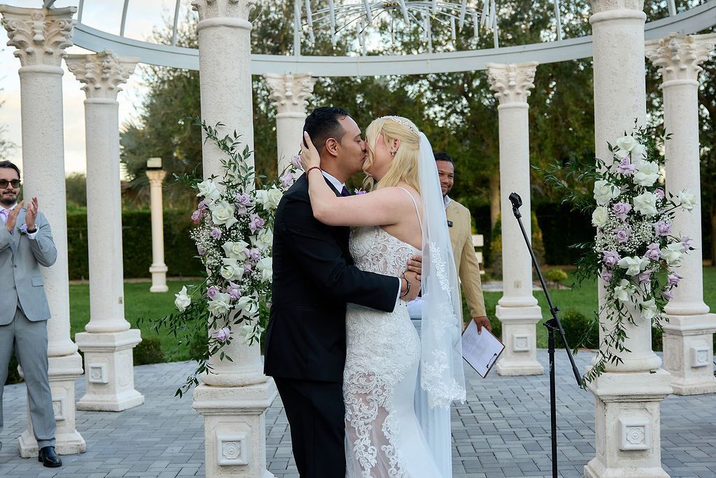 Newlyweds kissing under a gazebo decorated with flowers. Man in black suit, woman in white gown.